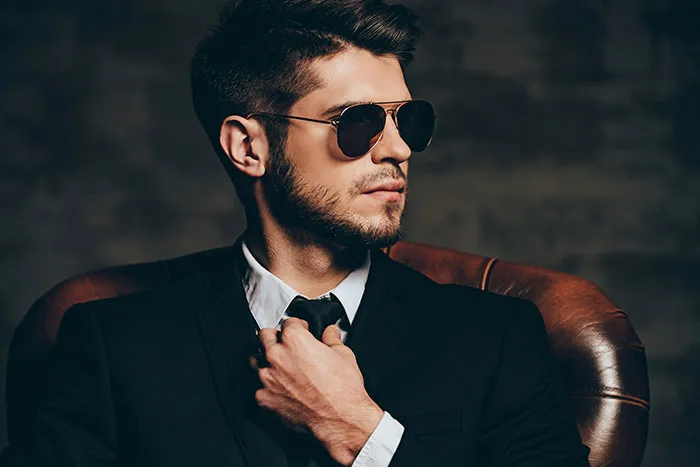 Man in a suit and sunglasses adjusting tie, sitting confidently, symbolizing out-of-touch wealthy lifestyle.
