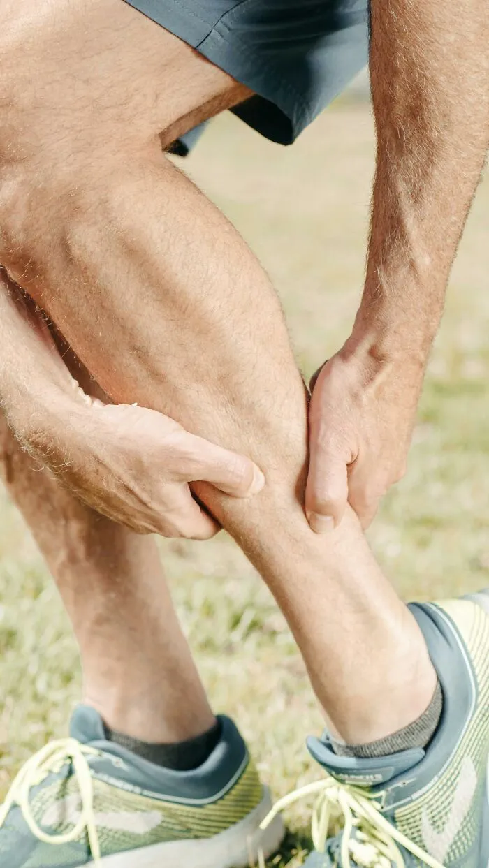 Person holds leg in concern, wearing athletic shoes outdoors.