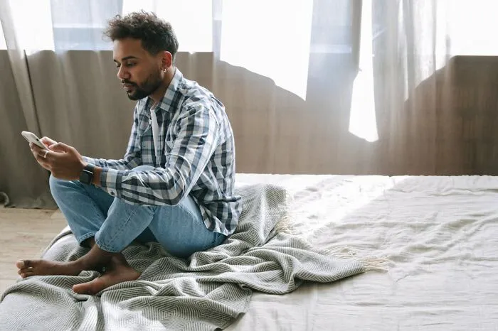 Man sitting on bed, using phone, amidst a tidy room, representing personal cleaning rules.