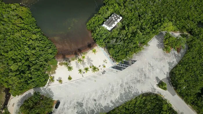Aerial view of an empty parking lot near a beach, surrounded by lush green trees.