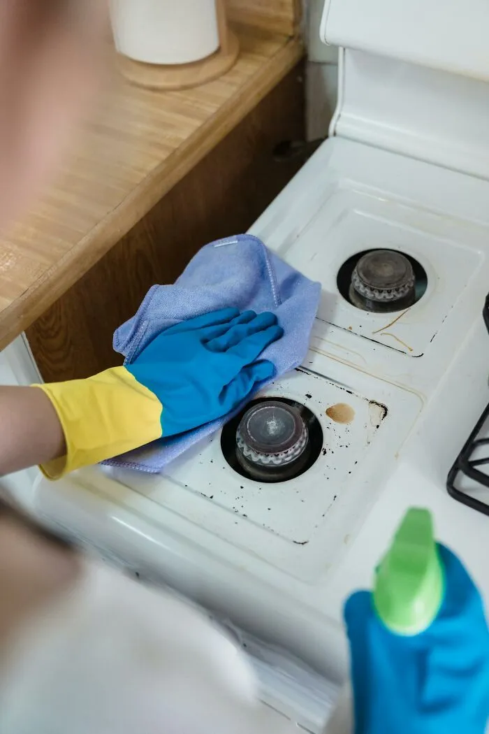 Person wearing gloves cleaning a stove top with spray and a cloth, illustrating strict cleaning rules.
