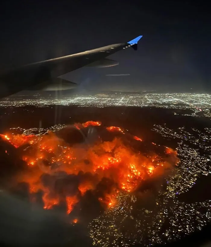 Aerial view from a plane of an intense wildfire illuminating a cityscape at night.