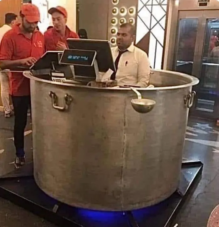 Man working inside a huge cooking pot with two staff members in red uniforms nearby in an unusual restaurant setup