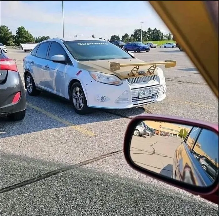 White car with a wooden spoiler attached to the hood in a parking lot, an image that might make you uncomfortable.