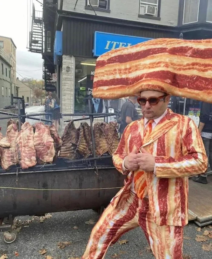 Man wearing a bacon suit and oversized bacon hat standing near a grill with ribs, creating an uncomfortable image.