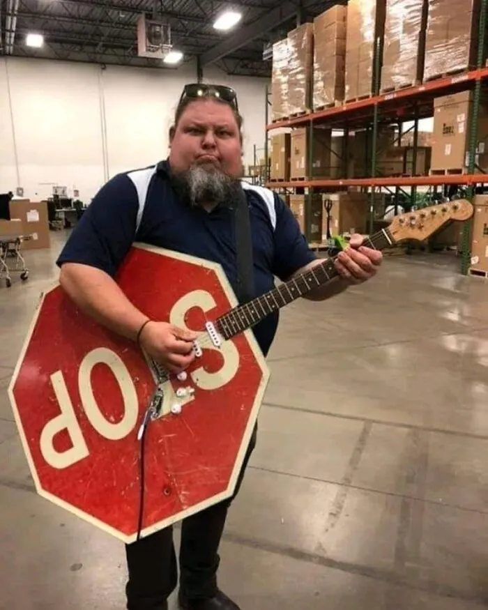 Man holding a guitar made from a stop sign, standing in a warehouse, an image that might make you uncomfortable.
