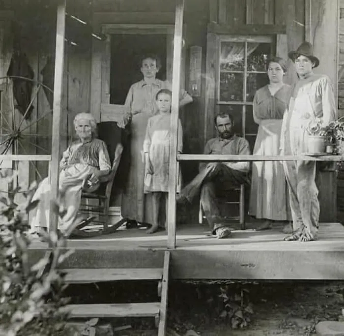 Black and white antique historical photograph showing a family gathered on a wooden porch in old-fashioned clothing.