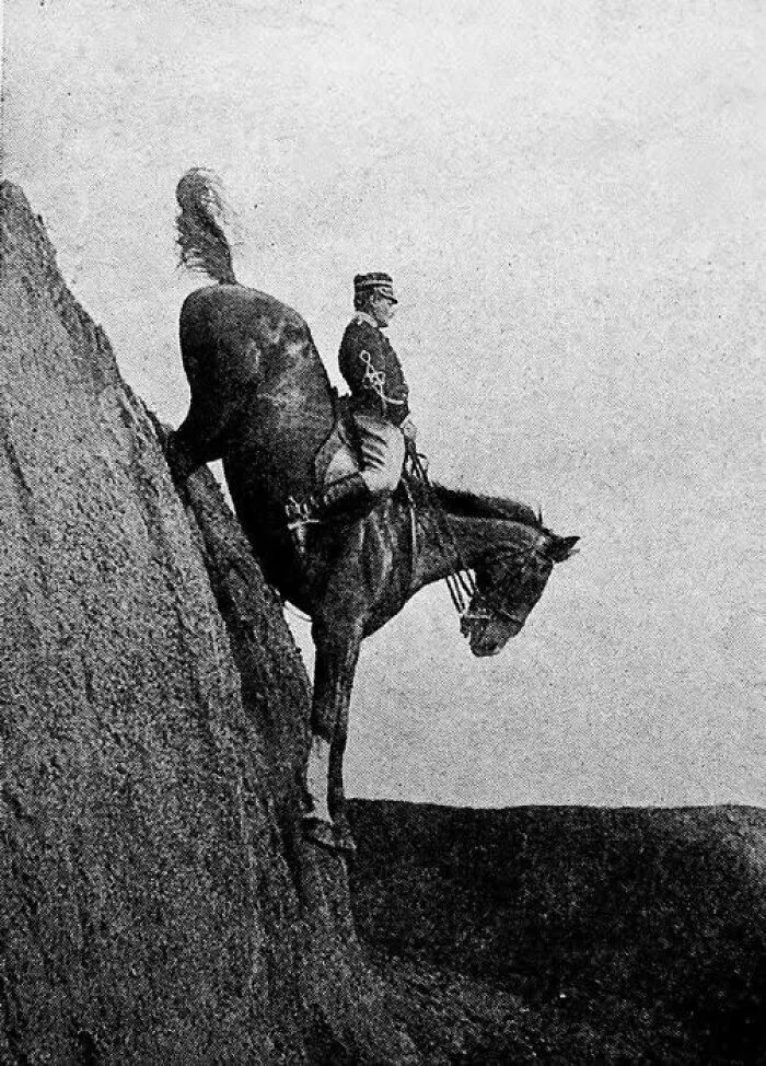 Antique historical photograph of a soldier riding a horse down a steep cliff in an old black and white image.