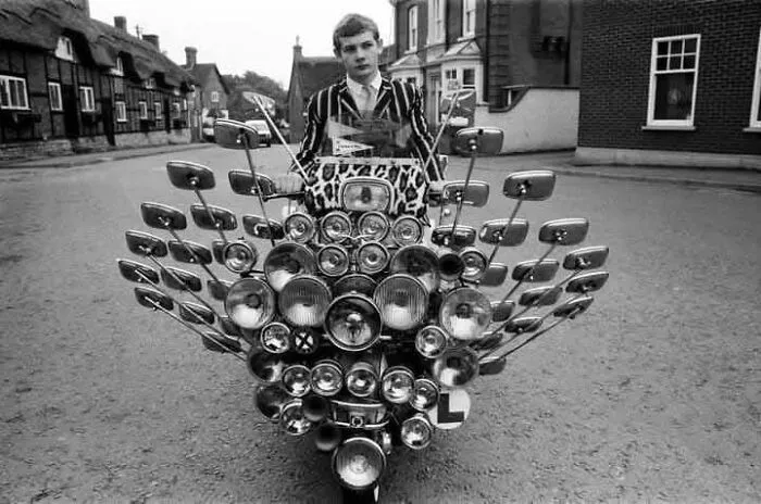 Young man on a vintage scooter with numerous mirrors and headlights in an antique historical photograph on a quiet street.