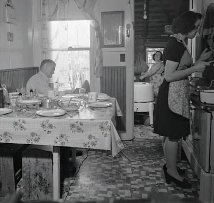 Older black and white antique historical photograph showing a woman cooking and people sitting in a vintage kitchen.