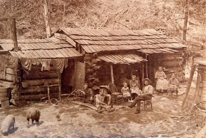 Family gathered outside a rustic log cabin in an antique historical photograph depicting life from the past.