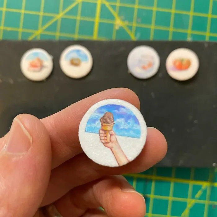 Hand holding a tiny one-inch painting of an ice cream cone against a blue sky with more miniature paintings blurred in the background.