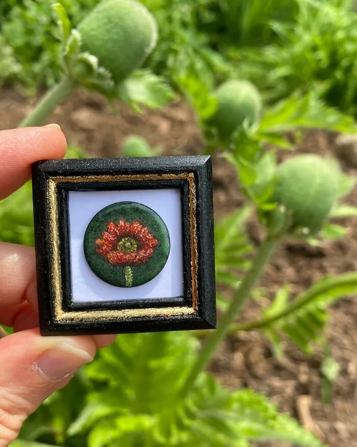Hand holding a tiny one-inch painting of a red flower in a black and gold frame outdoors with green plants in background