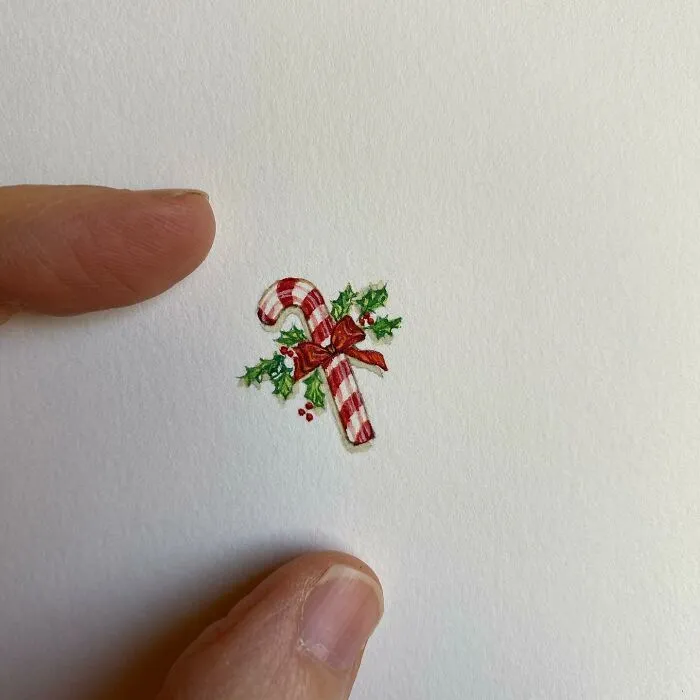 Tiny one-inch painting of a candy cane with holly, held between fingers, showcasing detailed everyday art by Brooke Rothshank.
