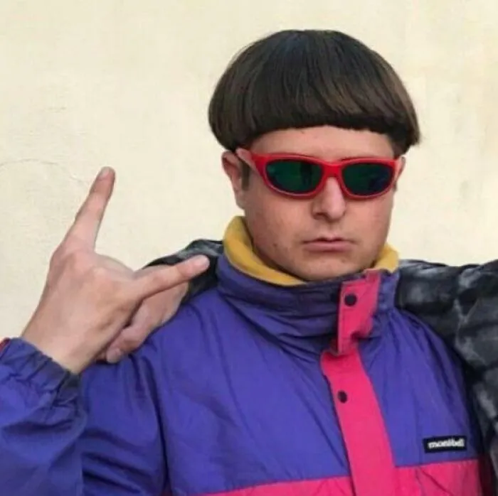 Person wearing cringey nineties fashion with bowl haircut, red sunglasses, and a colorful windbreaker jacket.