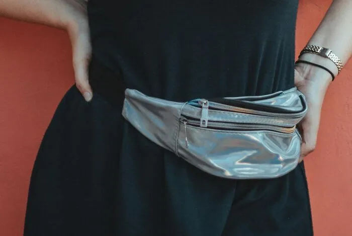 Person wearing a silver fanny pack around the waist, showcasing cringey nineties fashion accessories against an orange background.