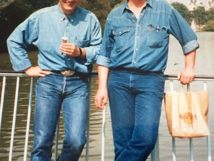 Two men wearing cringey nineties fashions in denim outfits, standing by a railing near a body of water outdoors.