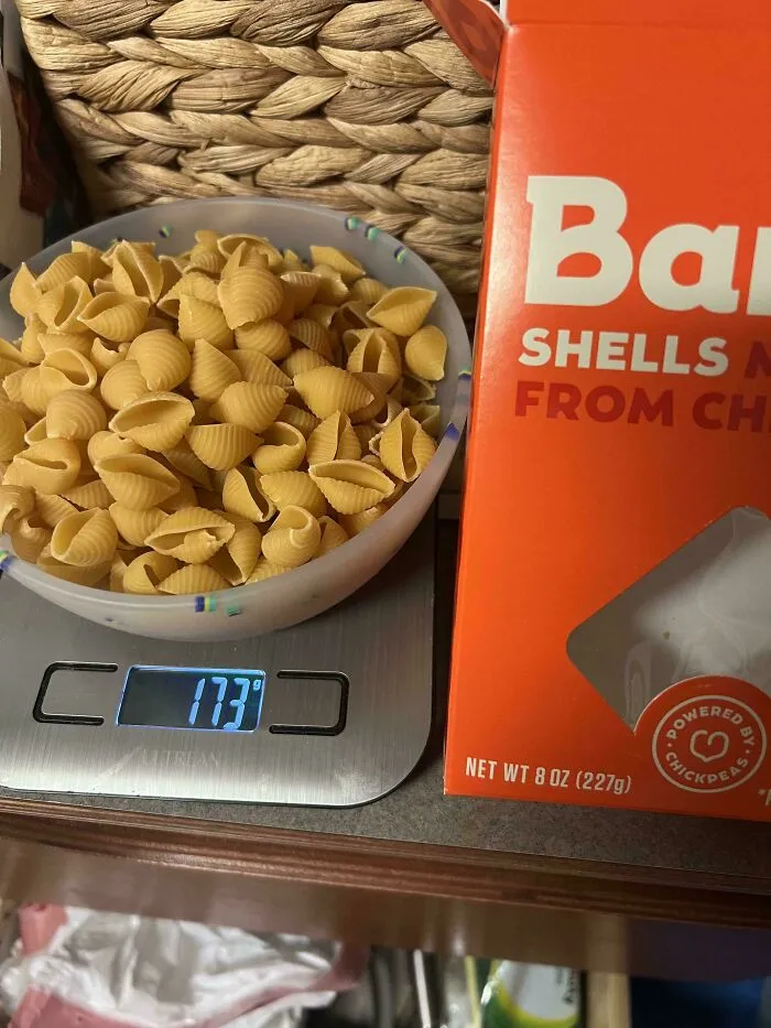 Bowl of pasta shells on a digital scale showing 173 grams beside a smaller, more expensive pasta product box.