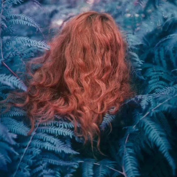 Woman with vibrant red hair surrounded by blue-toned foliage, captured in an intimate, cinematic-looking vintage camera photo.