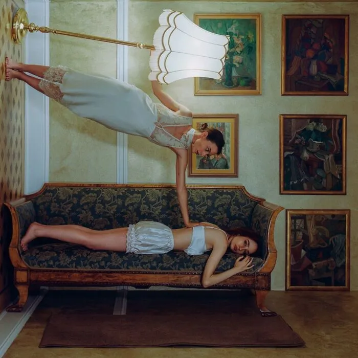 Two women in vintage-inspired outfits pose in a cinematic-looking shot, one balancing upside down holding a lamp, captured with vintage cameras.