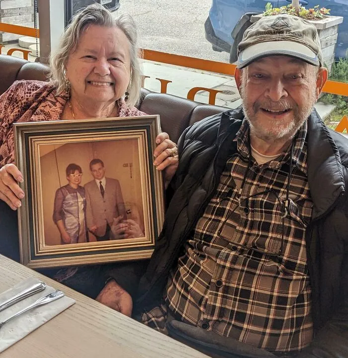 Elderly couple smiling at a restaurant holding a framed photo of their younger selves showing love unchanged by time.