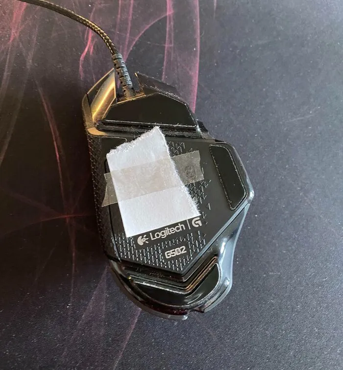 Wired Logitech G502 computer mouse with tape and paper on the bottom, showing a work-from-home setup humor.