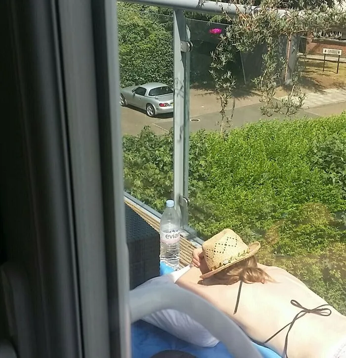 Person in a straw hat relaxing on a balcony during work-from-home, enjoying a sunny break with a water bottle nearby.