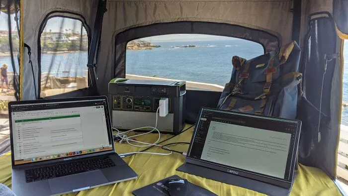 Portable work-from-home setup with two laptops and power station inside tent overlooking ocean view.