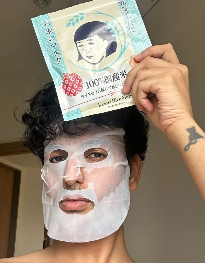 Person wearing a facial sheet mask while holding packaging, illustrating self-care in a work-from-home setting.