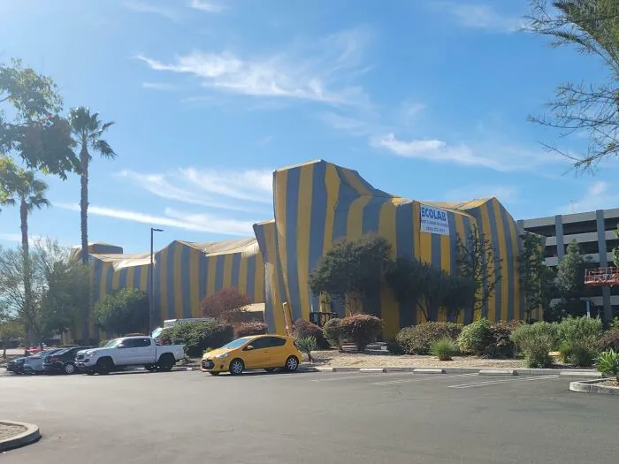 Edificio envuelto en telas amarillas y negras en un estacionamiento con autos y árboles bajo un cielo azul claro.
