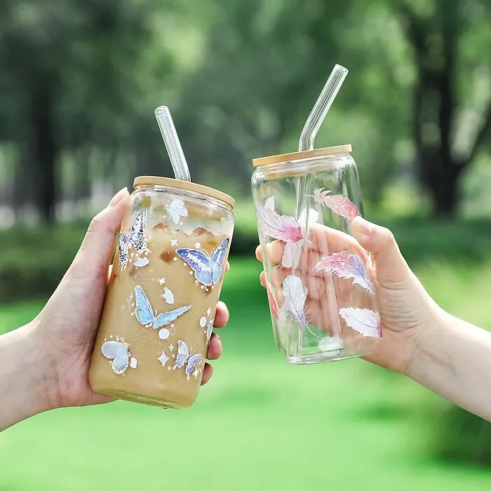 Two hands holding affordable Amazon finds of butterfly and feather glass tumblers with reusable straws outdoors.