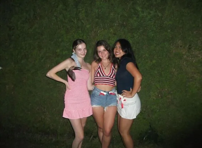 Three young women posing outdoors at night with a small animal on one woman’s shoulder in a casual wildlife moment.