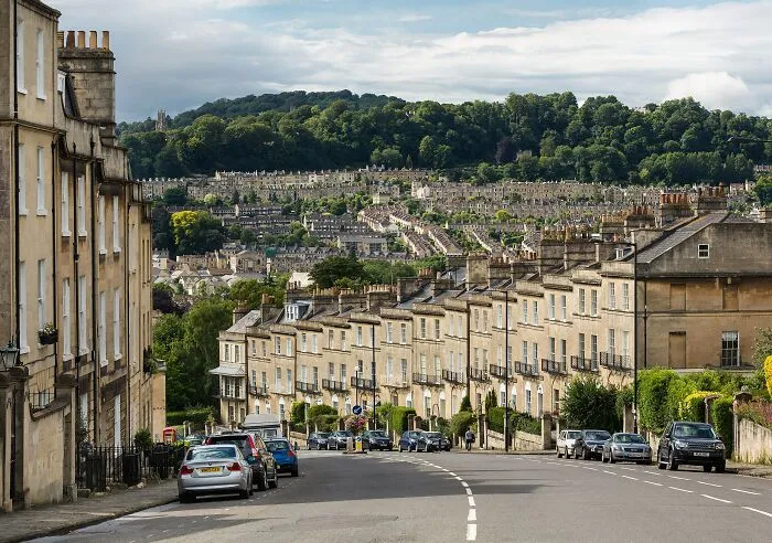 Historic row of stone buildings lining a street with cars, surrounded by trees and hills, showcasing historical artifacts ambiance.