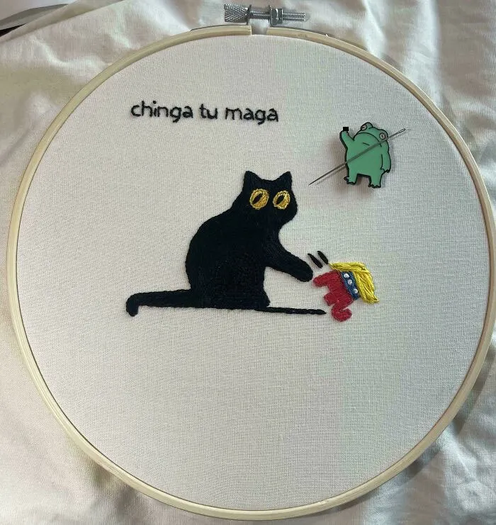 Black cat embroidery design with yellow eyes holding a small figure, displayed in an embroidery hoop on white fabric.