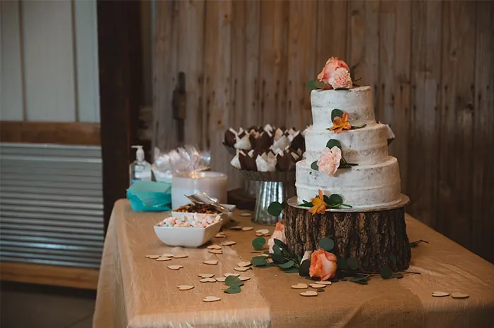 Pastel de bodas rústico con flores decorativas sobre base de madera en mesa con postres y decoración sencilla.