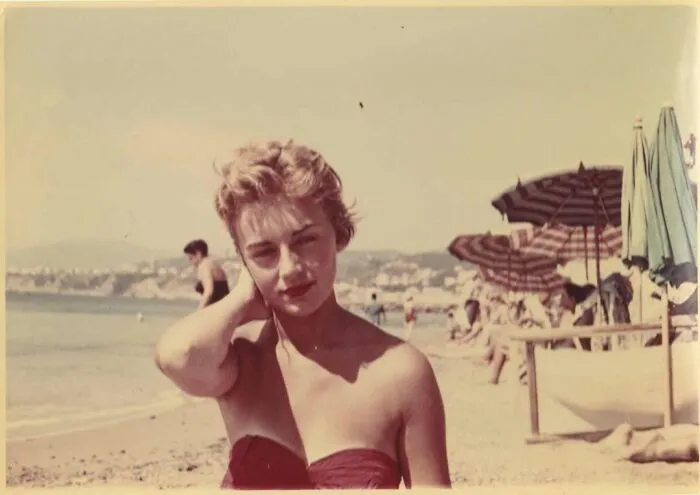 Vintage photo of a woman at the beach in a swimsuit with umbrellas and people in the background, capturing history.
