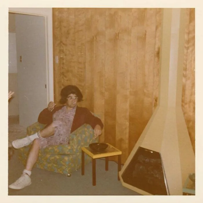 Vintage photo of a person sitting in a patterned armchair next to a retro fireplace in a wood-paneled room.