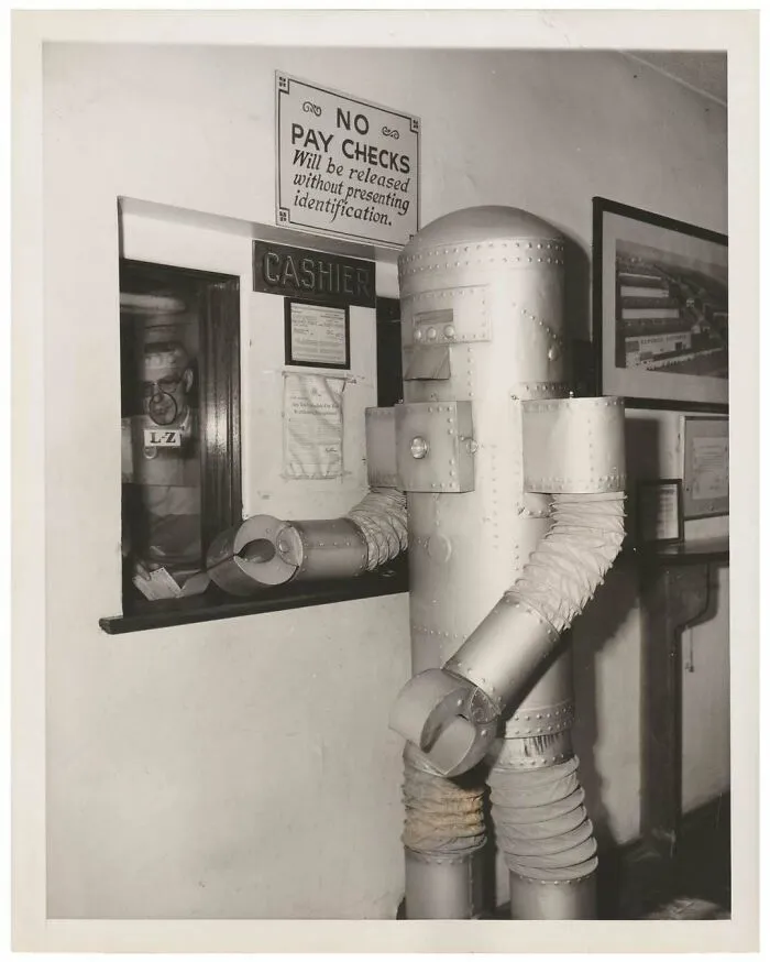 Vintage photo of a person in a homemade robot costume standing at a cashier window in an office setting.