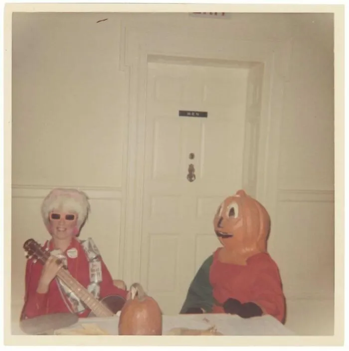 Vintage photo of two people in costumes, one playing guitar, beside a pumpkin on a table in front of a men’s restroom door.