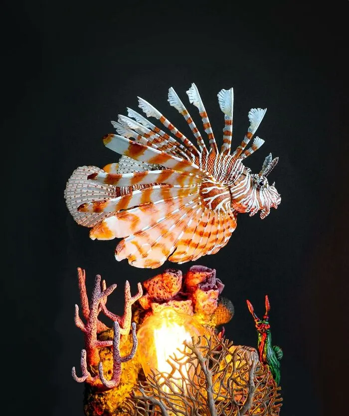 Lifelike bird sculpture crafted from paper and wood, displayed above coral art with glowing light on dark background.
