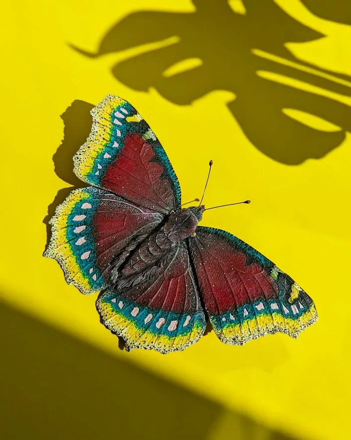 Lifelike butterfly crafted from paper and wood with detailed colorful wings, set against a bright yellow background.