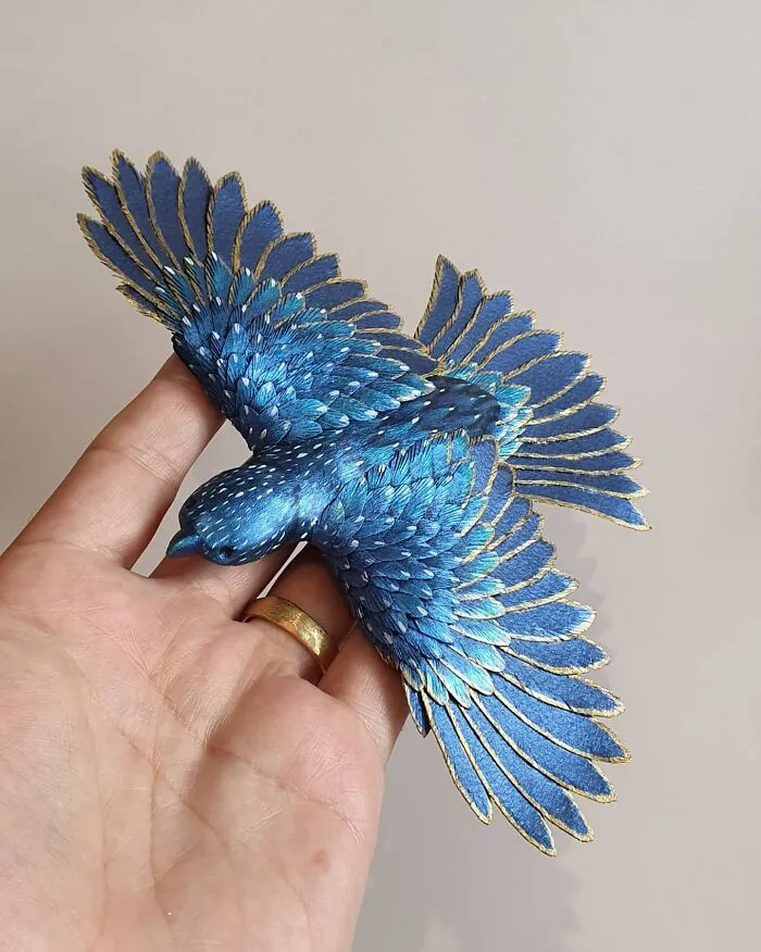 Hand holding a lifelike blue bird crafted from paper and wood with detailed feathers and gold edges.