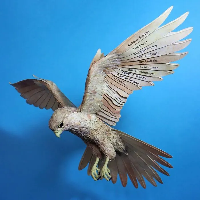 Lifelike bird sculpture crafted from paper and wood with detailed feathers and realistic form against a blue background.