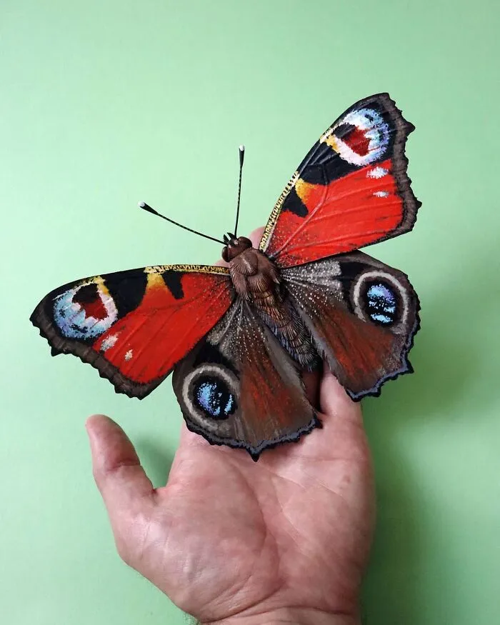 Hand holding a lifelike butterfly crafted from paper and wood, showcasing detailed and colorful wings against a green background.
