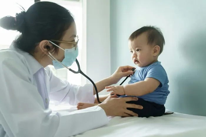 Pediatrician wearing mask using stethoscope to examine baby boy in blue shirt during a medical checkup session.