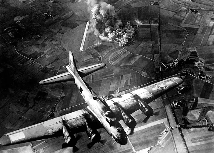 Black and white aerial photo of a bomber plane over farmland with smoke from an explosion in historical events.