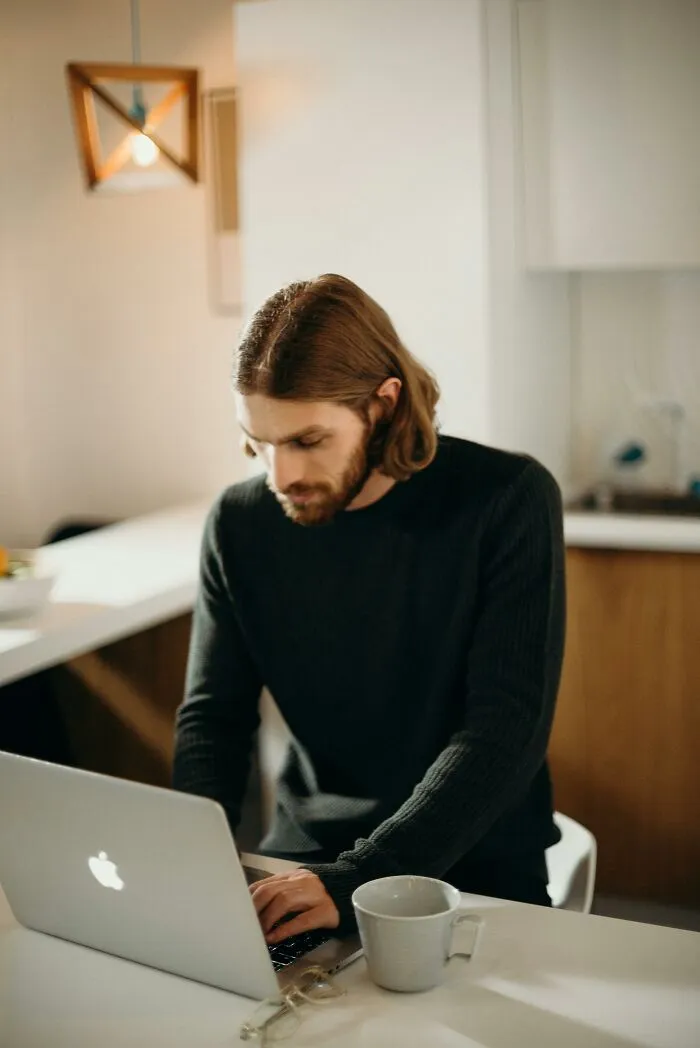 Man with long hair using a laptop in a kitchen, reflecting on out-of-touch comments that still provoke strong feelings.