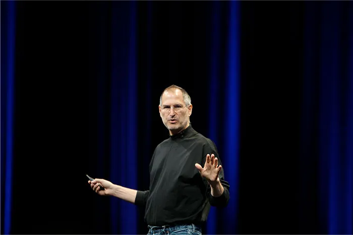 Steve Jobs speaking on stage wearing a black turtleneck and jeans during a presentation with blue curtains backdrop