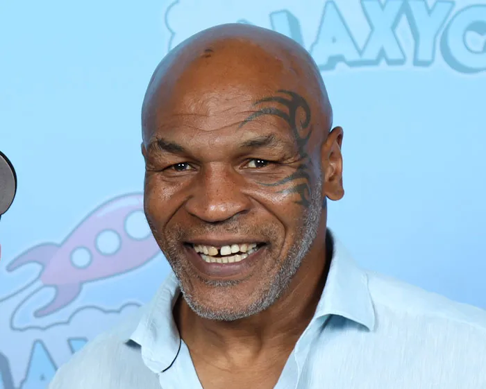 Smiling man with facial tattoo wearing a light blue shirt at an event with a blue background featuring logos.