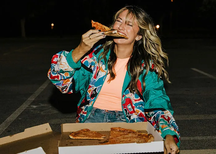 Young woman enjoying a slice of pizza at night, wearing colorful jacket, representing people who exposed cheating partners.